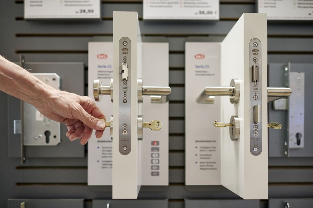 Die Hand einer Person steckt einen Schlüssel in das Schloss einer Ausstellungstür mit mehreren Schlössern. In einem Geschäft werden unterschiedliche Türschlossmechanismen vorgeführt.